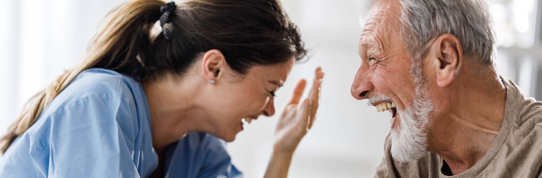 Helfende Altenpflegerin im vertraulichen Gespräch mit Bewohnerin, beide lachen.